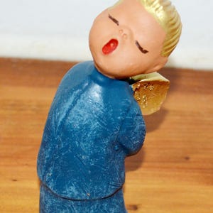 Puede incluir: Una pequeña figura de una persona con cabello rubio, ojos cerrados y boca abierta, que viste una túnica azul texturizada. La figura presenta un ala de color dorado en la espalda, una tez bronceada y el interior de la boca pintado de rojo.