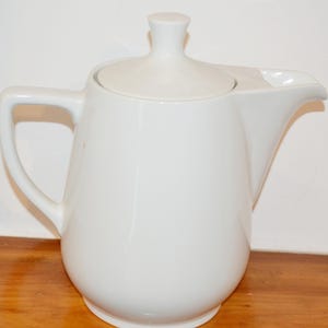 May include: A white ceramic teapot with a smooth, glossy finish, featuring a rounded body, a curved handle, a short spout, and a matching lid with a small knob handle on top. The design is simple and functional with clean, solid white lines.