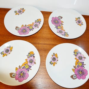 Könnte beinhalten: Ein Set aus vier runden weißen Keramiktellern, die jeweils mit einem Blumenmuster aus rosa, violetten und orangefarbenen Blüten und Blättern verziert sind, welche in einem geschwungenen Design auf der Oberfläche jedes Tellers angeordnet sind.