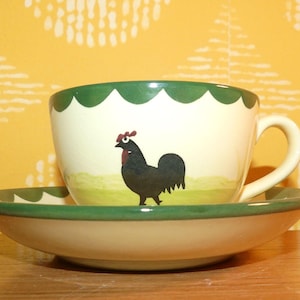 Peut inclure: Une tasse à thé et sa soucoupe en céramique crème, ornées d'un coq noir peint à la main avec une crête rouge sur la tasse. Le bord de la tasse et de la soucoupe présente une bordure verte festonnée, et la tasse possède une anse incurvée.