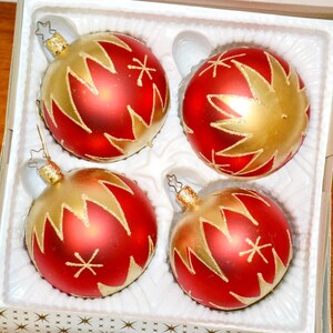 Könnte beinhalten: Ein Set aus vier kugelförmigen Weihnachtskugeln in einer weißen Kunststoffschale. Jede Kugel ist rot mit einem goldenen Sternmuster und Glitzerakzenten. Die Kugeln haben goldene Metallkappen mit Drahtschlaufen zum Aufhängen am Baum.
