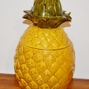 Peut inclure: Un pot en céramique en forme d'ananas avec un couvercle amovible. Le corps du pot est d'un jaune vif avec une surface texturée à motif en losange. Le couvercle présente un sommet vert brillant sculpté représentant les feuilles d'ananas.
