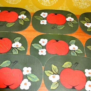 Pode incluir: Um conjunto de sete jogos americanos ovais com fundo verde escuro, uma ilustração central de uma maçã vermelha, folhas verdes e pequenas flores brancas. Cada peça apresenta um design botânico consistente com acabamento fosco.