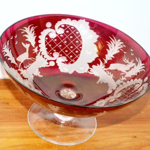 May include: A deep red cranberry glass bowl featuring intricate white etched designs of deer, foliage, and a lattice pattern. The bowl is supported by a clear glass pedestal base, showcasing a contrast between the coloured glass and clear stem.