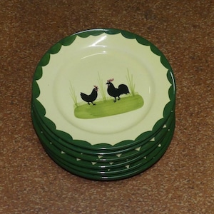 May include: A stack of five round ceramic plates with a cream-colored center and a dark green scalloped rim. The center of each plate features a green patch of grass with two black rooster silhouettes and thin green blades of grass.