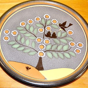 Puede incluir: Un plato redondo de cerámica con borde marrón oscuro y centro gris. El diseño presenta un árbol estilizado con hojas verde claro, flores blancas y naranjas, y varios pájaros negros posados en las ramas y en el suelo debajo.