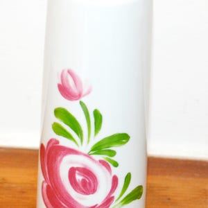 Könnte beinhalten: Eine zylindrische Vase aus weißem Porzellan mit einem handgemalten Motiv einer rosa Rose, grünen Blättern und einer kleinen rosa Knospe darüber. Die Vase hat eine glatte, glänzende Oberfläche und einen dünnen goldfarbenen Rand an der Öffnung.