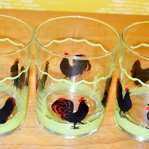 Peut inclure: Trois verres transparents ornés d'un motif répétitif de coq noir avec des accents rouges. Chaque verre présente une base peinte en vert clair et une ligne festonnée vert clair entourant la partie supérieure, formant une bordure décorative.
