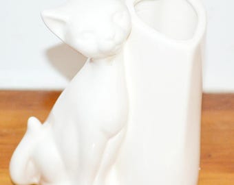 May include: A white ceramic vase featuring a sculpted cat figure attached to the side. The cat is depicted in a seated position with its head turned slightly. The vase opening has a heart-shaped rim, providing a distinct silhouette for this piece.