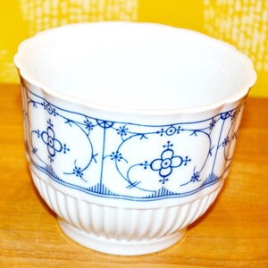 Könnte beinhalten: Eine weiße Porzellanschale mit gewelltem Rand und vertikal geripptem Boden. Die Außenseite ist mit einem wiederkehrenden blauen Blumenmuster verziert, das aus stilisierten vierblättrigen Blüten besteht, die durch feine, geschwungene Linien verbunden sind.