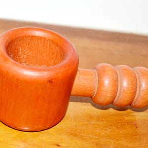 Könnte beinhalten: Ein zylindrischer Nussknacker aus Holz mit einer glatten, hellbraunen Oberfläche. Das Werkzeug verfügt über eine hohle Mitte zum Einlegen von Nüssen und einen horizontalen Griff mit drei ausgeprägten, abgerundeten, gerippten Abschnitten.