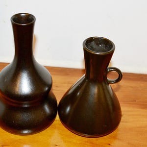 Könnte beinhalten: Zwei dunkelbraune Keramikvasen mit glatter, glänzender Oberfläche. Die linke Vase hat einen hohen, schlanken Hals und einen bauchigen, gestuften Körper. Die rechte Vase verfügt über einen breiteren, konischen Fuß und einen kleinen, runden Henkel.
