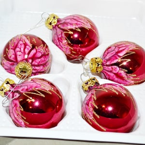Puede incluir: Un conjunto de cuatro adornos esféricos de vidrio con un acabado en rojo magenta intenso. Cada adorno presenta un patrón floral rosa pintado a mano con detalles de purpurina dorada y una tapa de metal dorado con un lazo de alambre plateado.