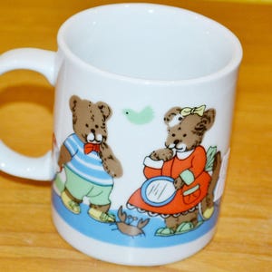 Può includere: Una tazza in ceramica bianca con un'illustrazione colorata di due orsacchiotti marroni. Un orso indossa una maglietta a righe blu e bianche con pantaloni verde chiaro, mentre l'altro indossa un vestito rosso con un fiocco giallo. Presenti un granchio e uccello.