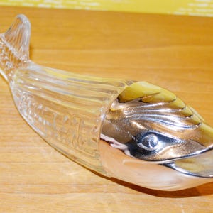 Puede incluir: Una botella de vidrio transparente con forma de pez que presenta una cabeza y una cola de color plateado metálico. El cuerpo del pez está hecho de vidrio transparente con detalles acanalados verticales, mientras que la cabeza es lisa y brillante.