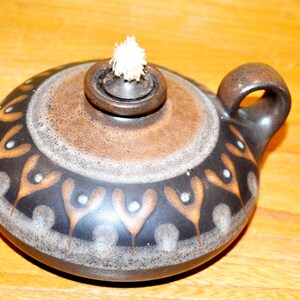 Op de afbeelding: Een ronde keramische olielamp met een donkerbruine en beige glazuurlaag. De lamp heeft een herhalend patroon van gestileerde oranje en witte bloemmotieven rondom het lichaam, een centrale metalen brander met een witte lont en een klein handvat.
