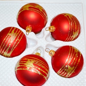 Puede incluir: Un conjunto de cuatro adornos navideños esféricos de color rojo con acabado mate, cada uno con un diseño de purpurina dorada de líneas horizontales y estrellas. Cada adorno tiene una tapa de metal dorado en la parte superior, dispuestos en una bandeja blanca.