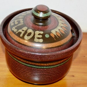 Könnte beinhalten: Ein runder, dunkelbrauner Keramik-Marmeladentopf mit passendem Deckel und Knaufgriff. Der Deckel ist mit dem Wort MARMELADE in heller Schrift, einem grünen Band und einem stilisierten Blattmuster verziert. Der Boden hat horizontale Rillen.