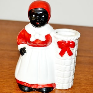 Puede incluir: Una figura de cerámica de una persona con un gorro rojo, una parte superior roja con cuello blanco y un delantal blanco, junto a un recipiente cilíndrico blanco con textura de cesta adornado con un lazo rojo. Tiene piel y zapatos negros.
