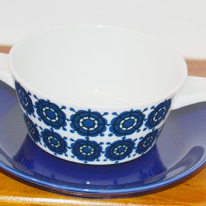May include: A white ceramic soup bowl with two side handles and a repeating blue and black circular geometric pattern, resting on a solid deep blue saucer. The bowl features a wide opening and a smooth finish, while the saucer is plain.
