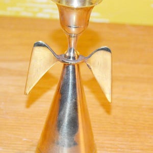 Puede incluir: Un candelabro de latón pulido con forma de ángel, que presenta una base cónica, dos alas planas extendidas y una copa circular ensanchada en la parte superior para sostener una vela. La superficie metálica es lisa y de tono dorado.