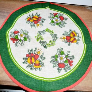 Puede incluir: Un mantel individual redondo de tela con centro color crema, borde verde y ribete rojo. El diseño incluye una corona verde central rodeada de motivos alternos de velas rojas con campanas amarillas y flores blancas con bayas rojas.