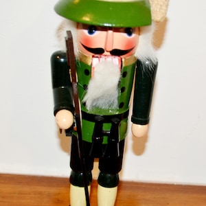 Könnte beinhalten: Eine hölzerne Nussknacker-Figur als Jäger verkleidet, mit grünem Hut, Tannenzapfen und Feder, grüner Jacke mit schwarzen Knöpfen, dunkler Hose und hellen Beinkleidern. Die Figur hat einen weißen Bart, einen Schnurrbart und hält ein Gewehr.