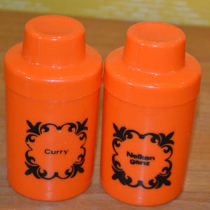 Peut inclure: Deux pots à épices cylindriques orange vif avec couvercles assortis. Chaque pot présente un cadre décoratif noir en filigrane imprimé sur le devant. Le pot de gauche est étiqueté Curry et celui de droite Nelken ganz en lettres noires.