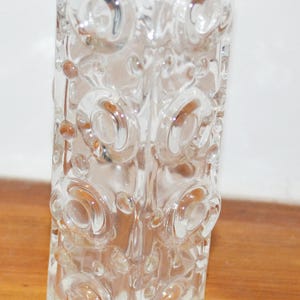 Puede incluir: Un florero de vidrio transparente con perfil cuadrado, que presenta un patrón repetitivo de motivos circulares en relieve y puntos pequeños grabados en sus cuatro lados. El recipiente tiene un centro hueco y una estructura gruesa y transparente.