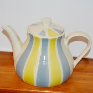 May include: A ceramic teapot featuring a glossy off-white base with vertical stripes in alternating shades of pale yellow and light blue. The teapot includes a matching lid with a small knob handle, a curved spout, and a rounded side handle.