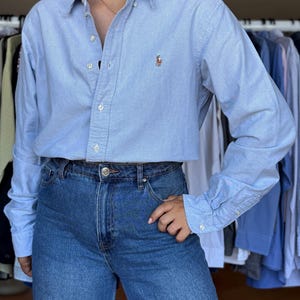 Könnte beinhalten: Ein hellblaues, langärmeliges Hemd mit Knopfleiste, klassischem Kragen und einem kleinen gestickten Logo auf der linken Brust. Das Hemd wird mit einer dunkelblauen Jeans mit hoher Taille und sichtbarem Metallknopfverschluss kombiniert.