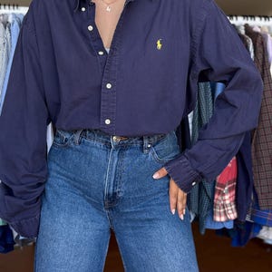 Könnte beinhalten: Ein langärmeliges, dunkelblaues Hemd mit Knopfleiste und einem gelben gestickten Logo auf der linken Brust, kombiniert mit einer blauen Jeans mit hoher Taille. Das Hemd hat einen klassischen Kragen, weiße Knöpfe und Manschetten, während die Jeans einen Reißverschluss und Knopfverschluss aufweist.