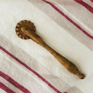May include: A wooden pastry wheel with a long, tapered handle and a circular, serrated cutting edge. The tool features a natural light brown wood grain finish with a smooth, turned handle design that ends in a small, rounded knob at the base.