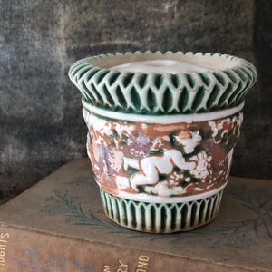 May include: A white ceramic planter with green and brown accents. The planter features a raised design of cherubs and grapes.