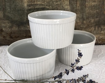 May include: Three white ceramic ramekins with fluted sides. The ramekins are stacked on top of each other. A sprig of lavender is in the foreground.