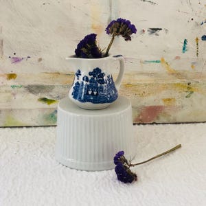 May include: A small, white and blue ceramic creamer with a handle, featuring a blue landscape design. The creamer sits atop a white, ribbed pedestal, and is adorned with dried purple flowers. The background is a textured, multi-colored surface.