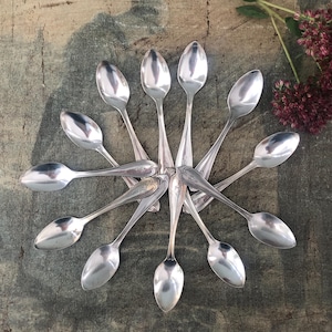 May include: A collection of eleven silver-toned metal spoons arranged in a circular sunburst pattern. Each spoon features a smooth, oval-shaped bowl and a slender handle, with some handles displaying a subtle engraved heart-shaped detail.