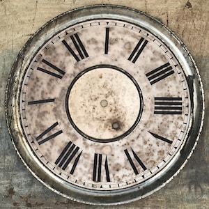 May include: Antique clock face with a distressed, aged appearance. The face is off-white with black Roman numerals and minute markers. A circular center is surrounded by a silver-toned metal rim. The clock face shows signs of wear and age.