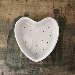 May include: A white ceramic heart-shaped dish featuring a series of small, evenly spaced drainage holes across the base. The interior surface is smooth and glossy, while the exterior rim is curved and defined, forming a distinct heart silhouette.