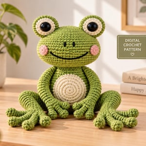 May include: A green crocheted frog toy with large black eyes, pink cheeks, and a cream-coloured belly. The frog has long limbs and webbed feet. A rectangular label on the right side features the text DIGITAL CROCHET PATTERN with a heart icon.