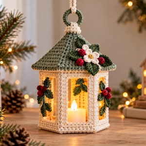 May include: A hexagonal crochet lantern featuring a cream-colored base and a sage green roof. The structure is decorated with crocheted holly leaves, red berries, and a white flower. A small, battery-operated white candle sits inside the lantern.