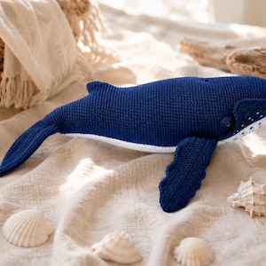 Op de afbeelding: Een donkerblauw gehaakte walvisknuffel met een witte onderbuik en een klein wit stippenpatroon bij de bek. De walvis heeft een textuur van garen, opvallende zijvinnen en een brede staartvin, vervaardigd met zichtbare steken.