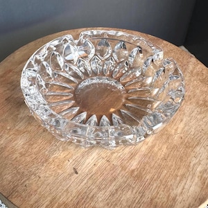 May include: A clear glass ashtray featuring a circular shape with a faceted, petal-like pattern radiating from the center toward the rim. The thick glass construction includes multiple notches along the outer edge for resting items.