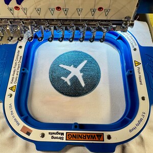 May include: A blue embroidery hoop holding white fabric with a circular teal patch featuring a white airplane silhouette. The hoop frame includes text warnings about strong magnets, pinch hazards, and instructions to keep away from computer disks.