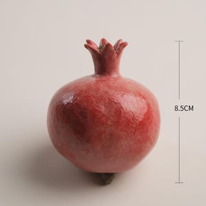 Op de afbeelding: Een rode granaatappelsculptuur met een glanzend, getextureerd oppervlak en een prominente kelk aan de bovenkant. Het object wordt getoond met maatlijnen die een hoogte van 8,5 cm en een breedte van 6 cm aangeven voor een duidelijke schaal.