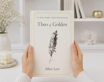 May include: A hardcover book titled Theo of Golden by Allen Levi with a cream-colored cover featuring a black ink illustration of a single feather in the center and the text #1 NEW YORK TIMES BESTSELLER printed at the top of the front cover.