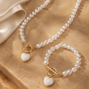 Puede incluir: Un conjunto de collar y pulsera con perlas blancas artificiales. Cada pieza incluye un cierre de barra dorado y un colgante liso en forma de corazón blanco con un borde dorado, creando un conjunto de joyería a juego.