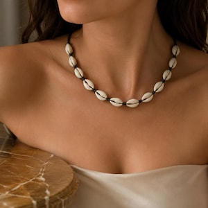 Puede incluir: Un collar que presenta una serie de conchas de cauri de color blanco roto y forma ovalada, ensartadas en un cordón fino de color marrón oscuro. Las conchas están espaciadas uniformemente alrededor del cuello, separadas por un nudo.
