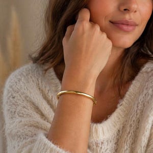 Puede incluir: Un brazalete rígido de tono dorado pulido con una superficie lisa y redondeada y un diseño circular continuo. El brazalete presenta un acabado metálico reflectante y una estética sencilla y sin adornos que rodea la muñeca de forma uniforme.