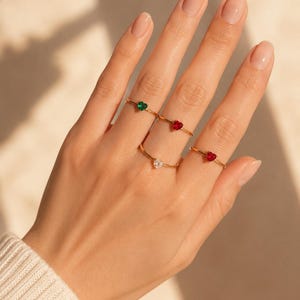 Puede incluir: Cuatro anillos finos de oro, cada uno con una pequeña piedra preciosa en forma de corazón. Los anillos incluyen uno con piedra verde, dos con piedras rojas y uno con piedra blanca transparente, lucidos en los dedos de una mano manicurada.